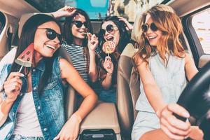 Great start of their journey. Four beautiful young cheerful women looking at each other with smile and holding lollipops while sitting in car photo