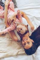 Morning hugs. Top view of cheerful beautiful young woman looking at camera with smile and hugging her baby girl while lying in bed photo
