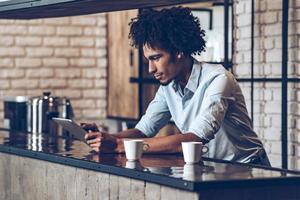 Running business easier with technologies. Side view of young African man using his digital tablet while leaning at bar counter with two coffee cups photo