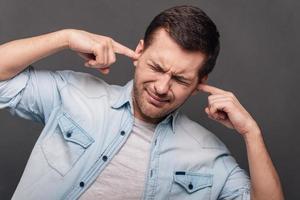 Too loud Young handsome man covering ears by hands and keeping eyes closed while standing against grey background photo