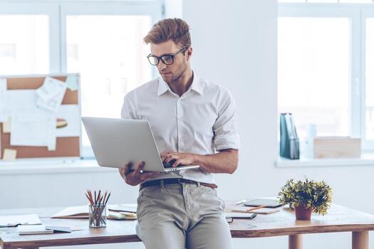 Managing everything with one click. Handsome young man in glasses using his laptop while leaning to the table in office photo