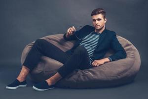 Stylish and handsome. Confident young handsome man holding his sunglasses and looking at camera while sitting on beanbag against grey background photo