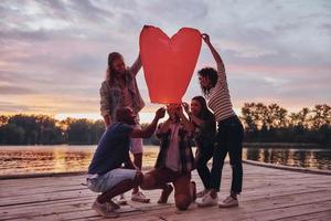Heart shaped lantern. Full length of young people in casual wear lighting up sky lantern while standing on the pier photo
