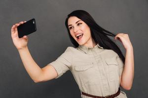 Capturing fun. Beautiful young woman making selfie by smart phone and smiling while standing against grey background photo