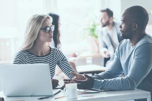 Discussing some business issues. Two young business people discussing something while sitting at the office desk together while their colleagues sitting in the background photo
