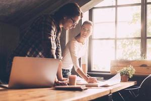 Finding solution together. Confident young man and woman in casual wear pointing blueprint and communicating while both standing near wooden desk in creative office photo
