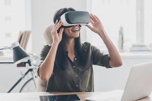 Future is right now. Confident young woman adjusting her virtual reality headset and smiling while sitting at her working place in office photo