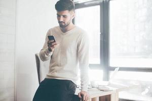 Typing quick message. Confident young man holding smart phone and looking at it while standing near his working place in office photo