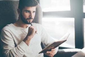 Waiting for inspiration. Thoughtful young man holding notebook and touching his chin with pen while sitting at his working place in office photo