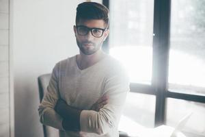 Confident business expert. Confident young man in eyeglasses keeping arms crossed and looking at camera while leaning at the desk in office photo