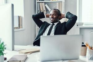 Happy young African man in formalwear smiling and keeping hands behind head while working in the office photo