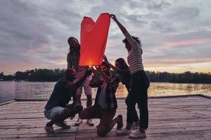 Heart shaped lantern. Full length of young people in casual wear lighting up sky lantern while standing on the pier photo