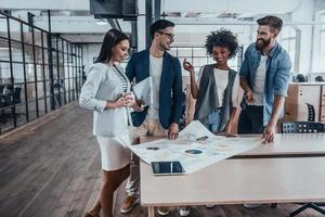 Best team ever. Group of young business people in smart casual wear working together and smiling while standing near the wooden desk in office photo