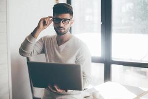 How may I help you  Confident young man holding laptop and adjusting his eyeglasses while leaning at the desk in office and looking at camera photo
