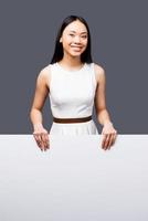 Fresh face for a new project. Beautiful young Asian woman looking at camera, holding hands on copy space and smiling while standing against grey background photo