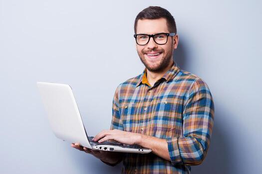 Working on laptop. Handsome young man working on laptop and smiling while standing against grey background photo