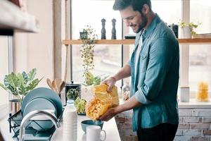 Easy breakfast. Handsome young man in casual wear preparing corn flakes and smiling while standing in the kitchen at home photo