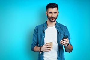 Handsome young man smiling and using smart phone while standing against blue background photo