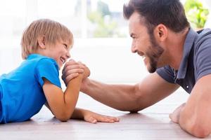 pequeño campeón. vista lateral de feliz padre e hijo compitiendo en la lucha de brazos mientras ambos yacen en el suelo de madera dura foto