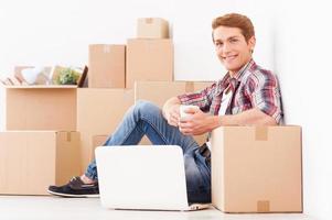 acaba de mudarse. vista lateral de un joven alegre sentado en el suelo de su nuevo apartamento y tomando café mientras cajas de cartón yacen en el fondo foto