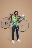 Full length of young African man in casual clothing looking at camera and carrying bicycle while standing against brown background photo
