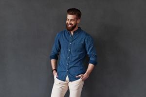 Too good to be real. Good looking young man keeping hands in pockets and smiling while standing against grey background photo