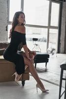 Feeling free and relaxed. Attractive young woman in elegant black dress with a deep slit posing while sitting on the sofa photo