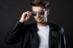 Cool and handsome. Handsome young man adjusting his sunglasses and looking at camera while standing against black background photo