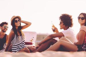 Spending nice time with friends. Cheerful young people spending nice time together while sitting on the beach and drinking beer photo