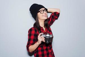 le gusta disparar. bella mujer joven con sombreros y anteojos sosteniendo la cámara y manteniendo los ojos cerrados mientras se enfrenta a un fondo gris foto