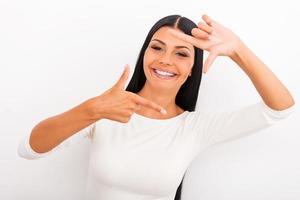 Gesturing finger frame. Cheerful young woman looking at camera and gesturing finger frame while standing against white background photo