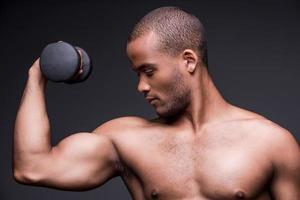 Exercising with dumbbell. Young shirtless African man exercising with dumbbell while standing against grey background photo