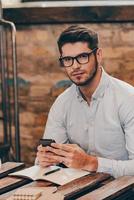 Always in touch.Handsome thoughtful young man holding smart phone and looking at camera while sitting at his working place photo