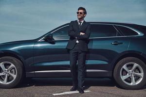 Confident in his style. Full length of handsome young businessman keeping arms crossed and smiling while standing near his car outdoors photo