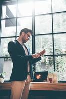 Typing quick message to colleague. Low angle view of confident young man holding smart phone and looking at it while standing in front of the big window in office or cafe photo