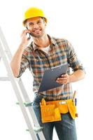 reparador alegre. joven trabajador manual sonriente hablando por teléfono móvil y sosteniendo el portapapeles mientras se inclina en la escalera foto