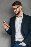 Great news Confident young man in glasses using his smartphone with smile and keeping hand in pocket while standing against grey background photo