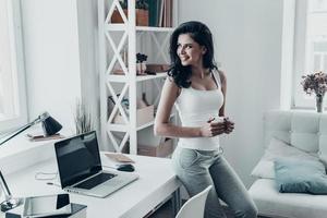 de buen humor. hermosa joven mirando afuera y sosteniendo una taza mientras se apoya en el escritorio en casa foto