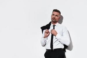 Successful professional. Handsome young man in full suit carrying jacket on shoulders and looking away with smile while standing against white background photo