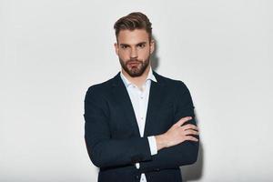 Confident in his perfect style. Handsome young man in full suit keeping arms crossed and looking at camera while standing against grey background photo