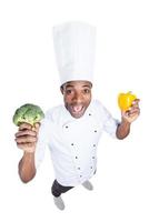 Cooking healthy food. Top view of excited young African chef in white uniform stretching out broccoli and pepper while standing against white background photo