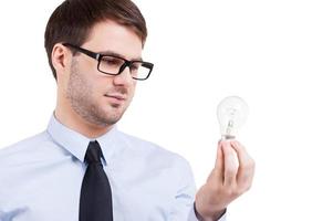 In search of new ideas. Thoughtful young man in shirt and tie holding lighting bulb and looking at it while standing isolated on white photo