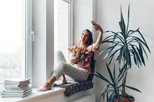 Happy young woman in cozy pajamas taking selfie with her bouquet of tulips while resting on the window sill at home photo