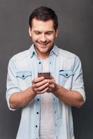 Great message Cheerful young man using his smartphone with smile while standing against grey background photo