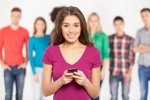 Always in touch. Cheerful young woman holding mobile phone and smiling while his friends standing on background photo