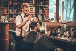 Making hair look magical. Young bearded man getting haircut by hairdresser while sitting in chair at barbershop photo