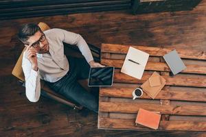 Confidence and success. Top view of handsome young man holding touchpad and looking at camera while sitting at his working place photo