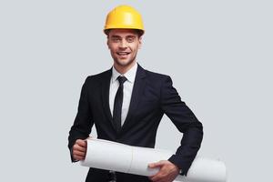 Achieving best results. Good looking young man in hardhat carrying blueprint and smiling while standing against grey background photo