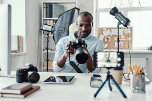 Charming young African man in shirt showing digital camera and telling something while making social media video photo