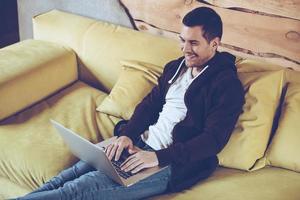 Surfing the net. Top view of cheerful young man using his laptop with smile while sitting on couch at home photo